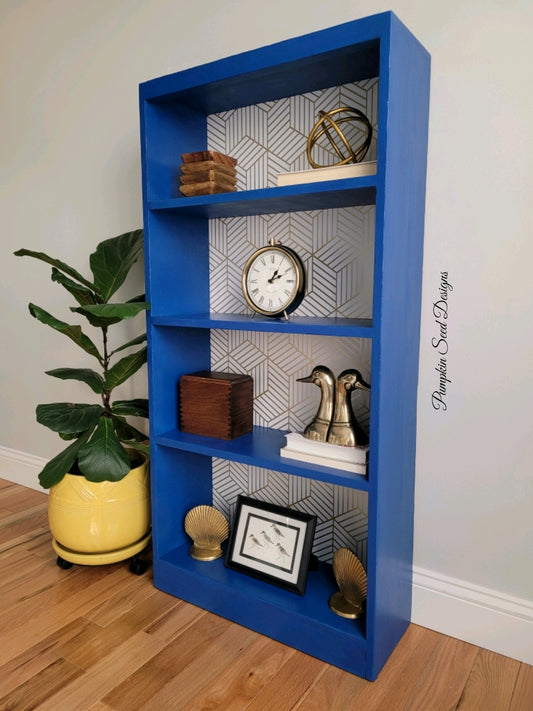 Royal blue painted bookshelf with white and gold geometric patterned wallpaper backing next to yellow plant pot