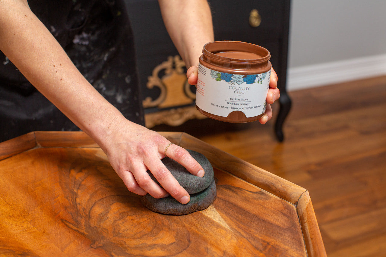 A Country Chic Paint Painting Sponge applying brown furniture glaze on a wood table top